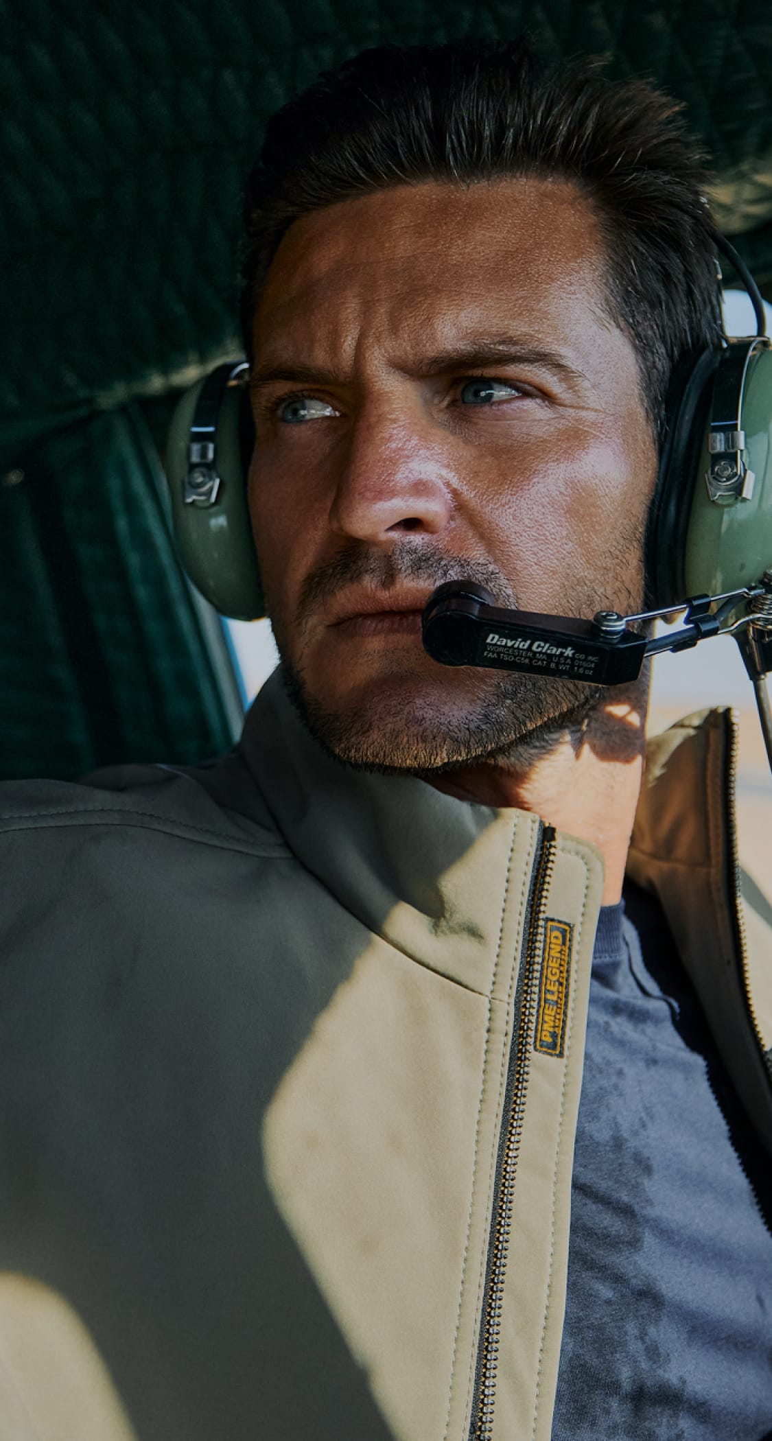 Man wearing a headset with a microphone, looking out of a window. He is dressed in a light jacket and appears focused.