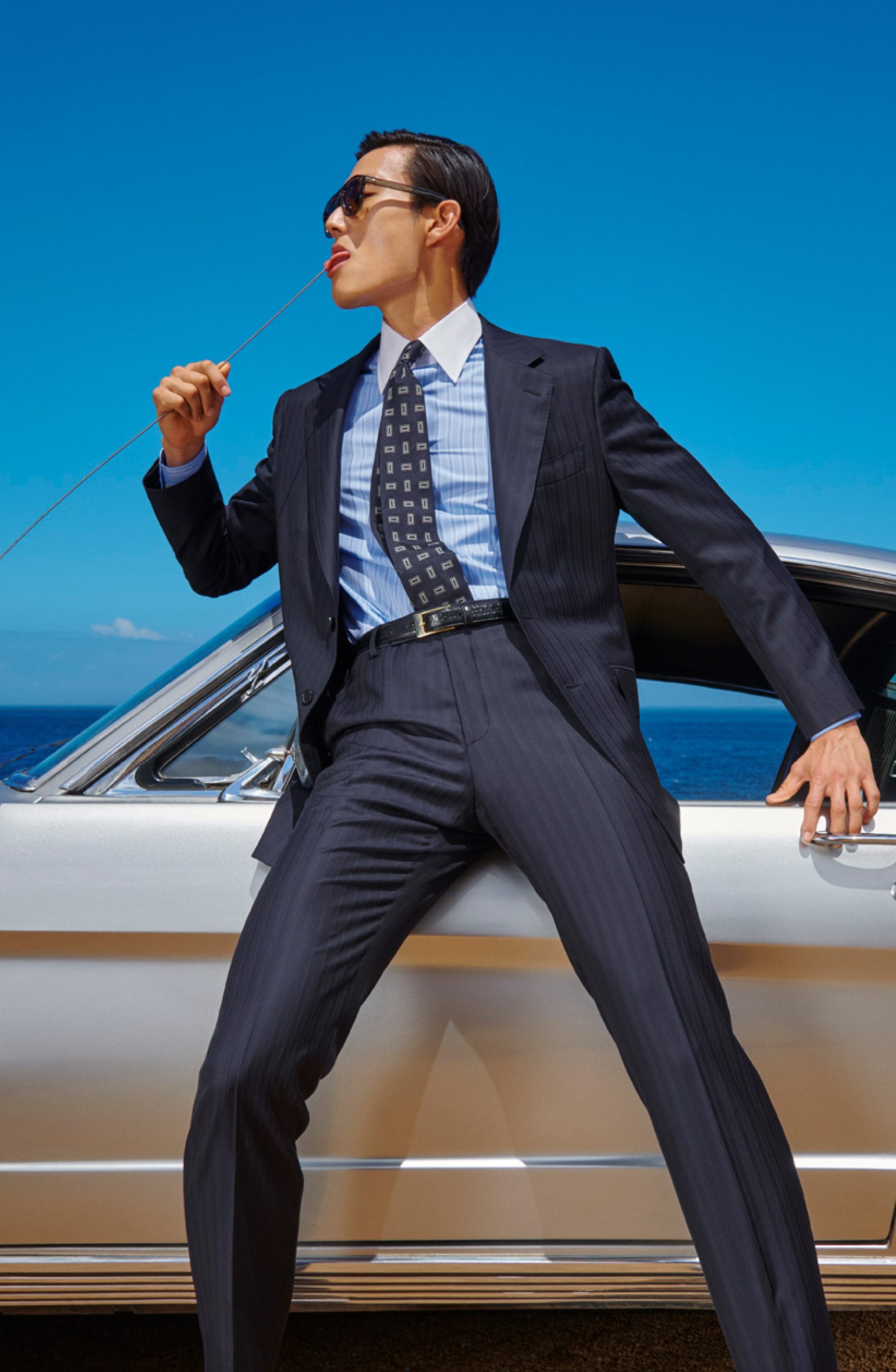 Man in a suit and sunglasses leans against a car, playfully holding a string with his mouth against a clear blue sky.