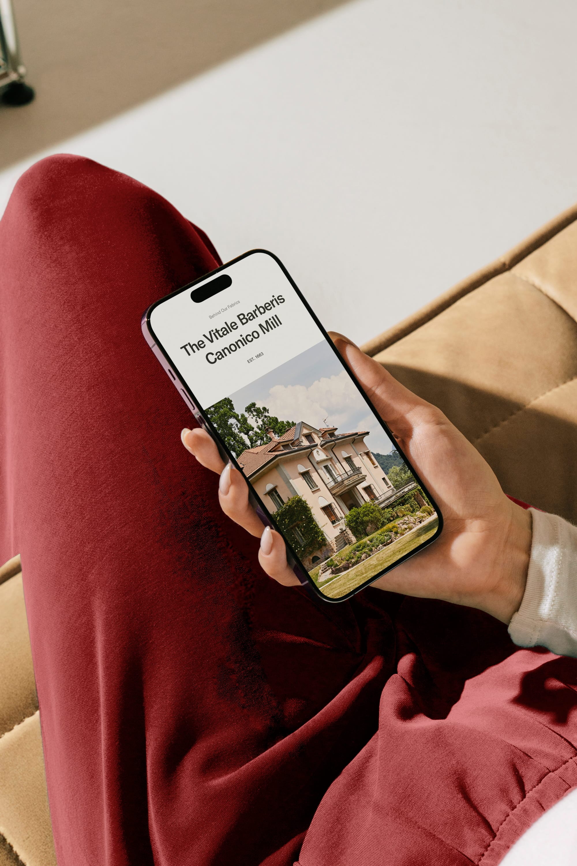 Person holding a smartphone displaying "The Vitale Barberis Canonico Mill" with building image; wearing red pants and sitting on a beige surface.
