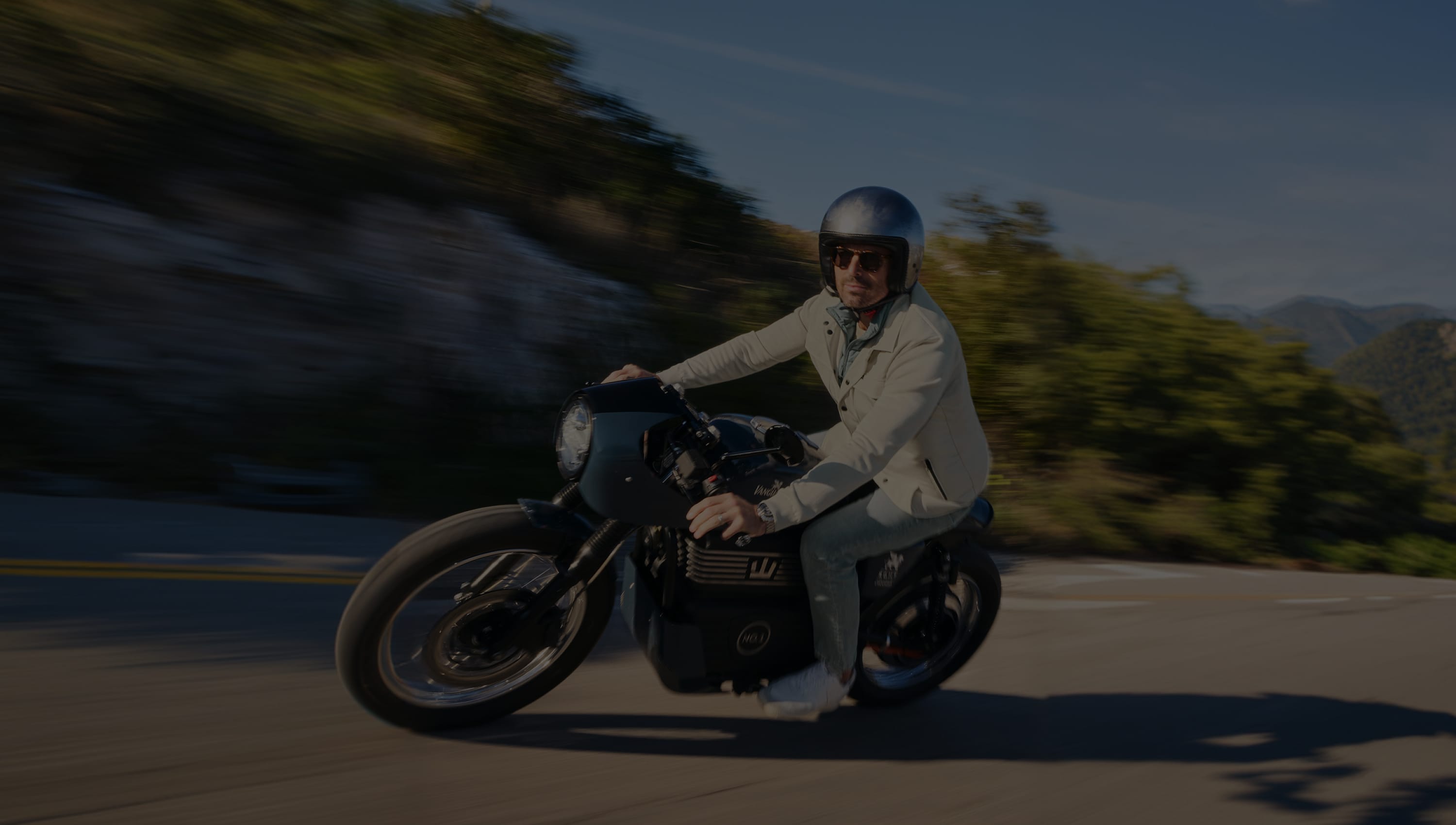 A man in a light jacket and helmet rides a motorcycle on a scenic, winding road with blurred greenery in the background.