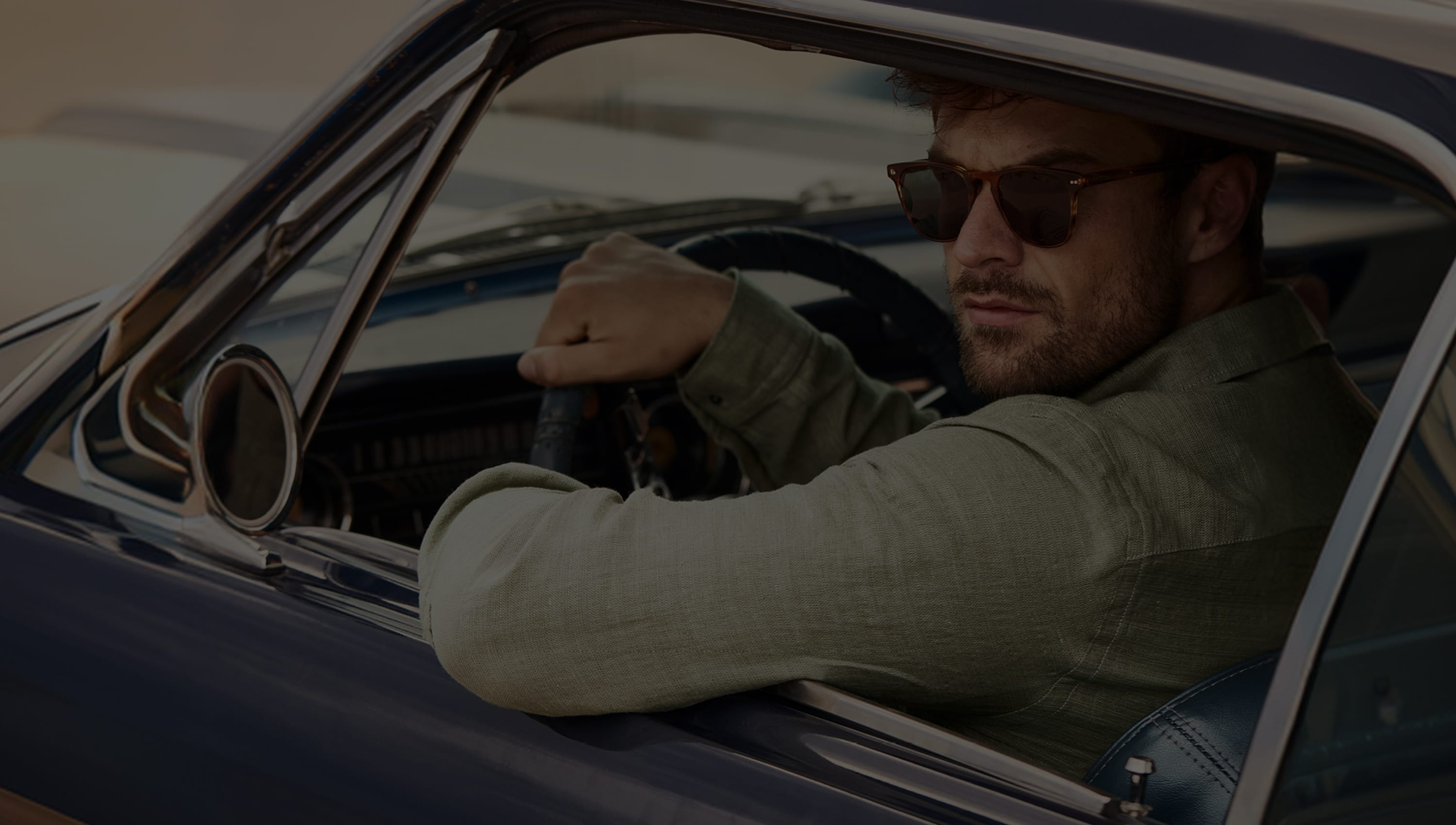 Man wearing sunglasses and a green shirt sitting in a classic car, looking out the open window with a focused expression.