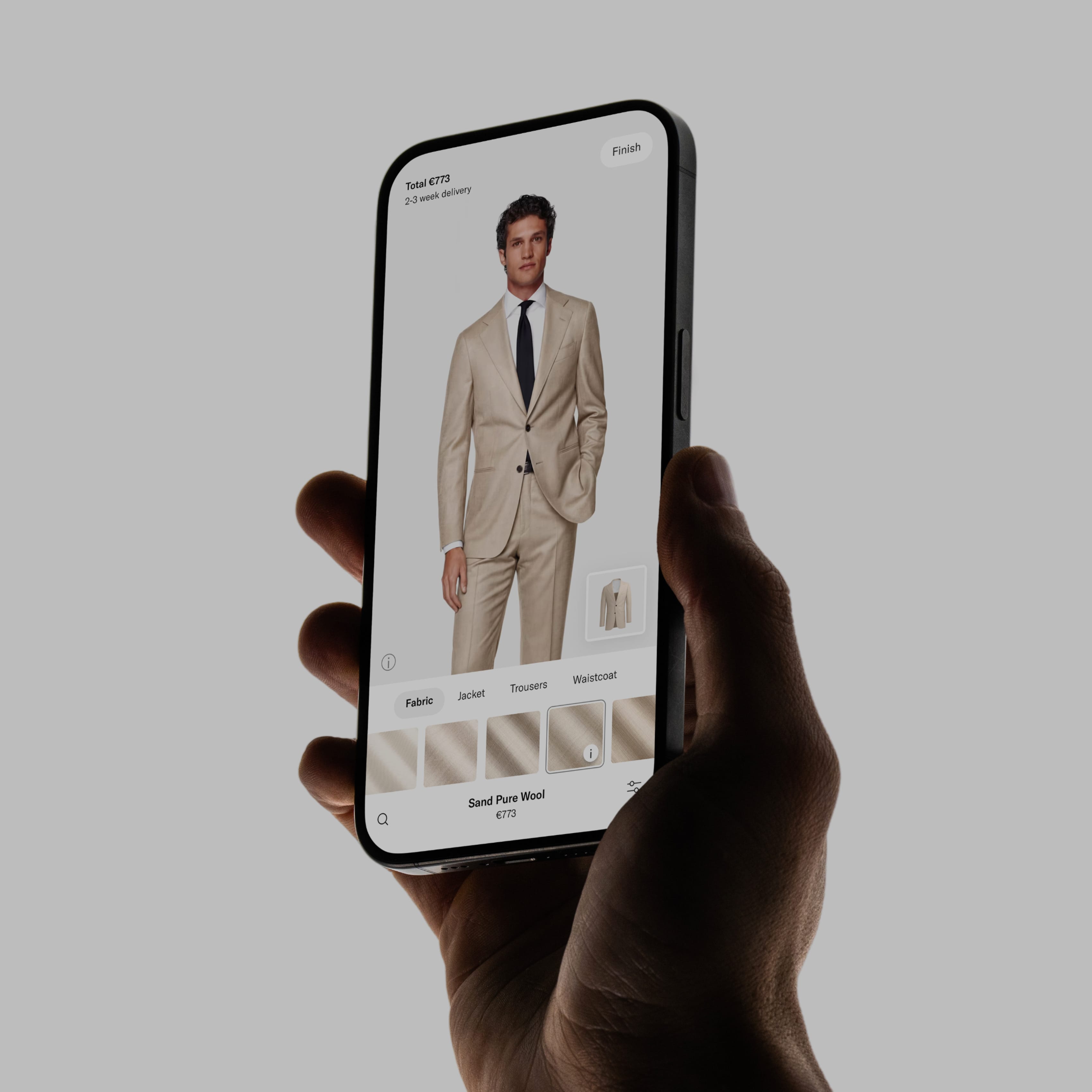Hand holding a smartphone showing a beige suit selection on a shopping app, with fabric and style options visible.