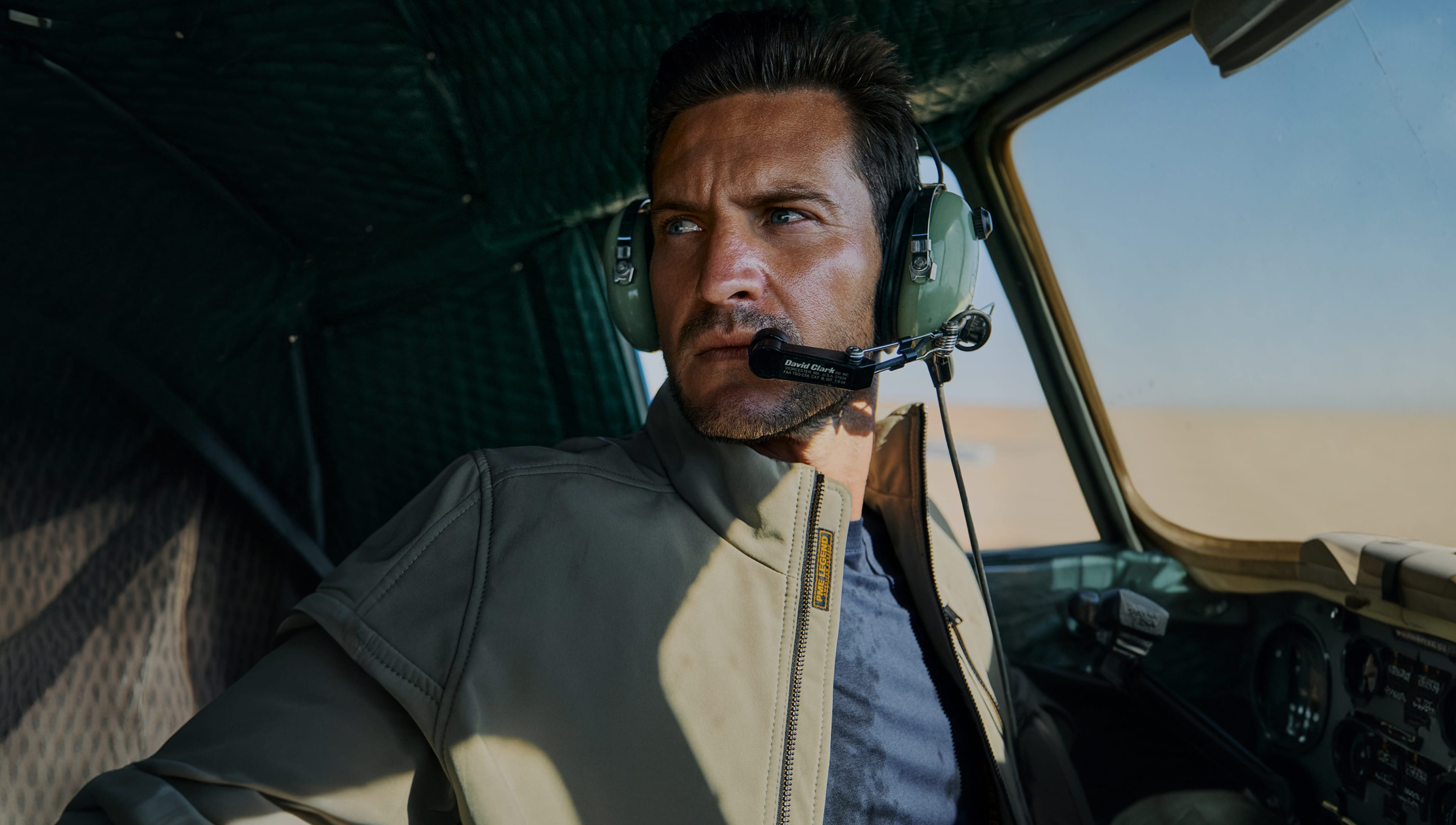 Man in a cockpit wearing a headset, looking out the window with a focused expression. Desert landscape visible outside.