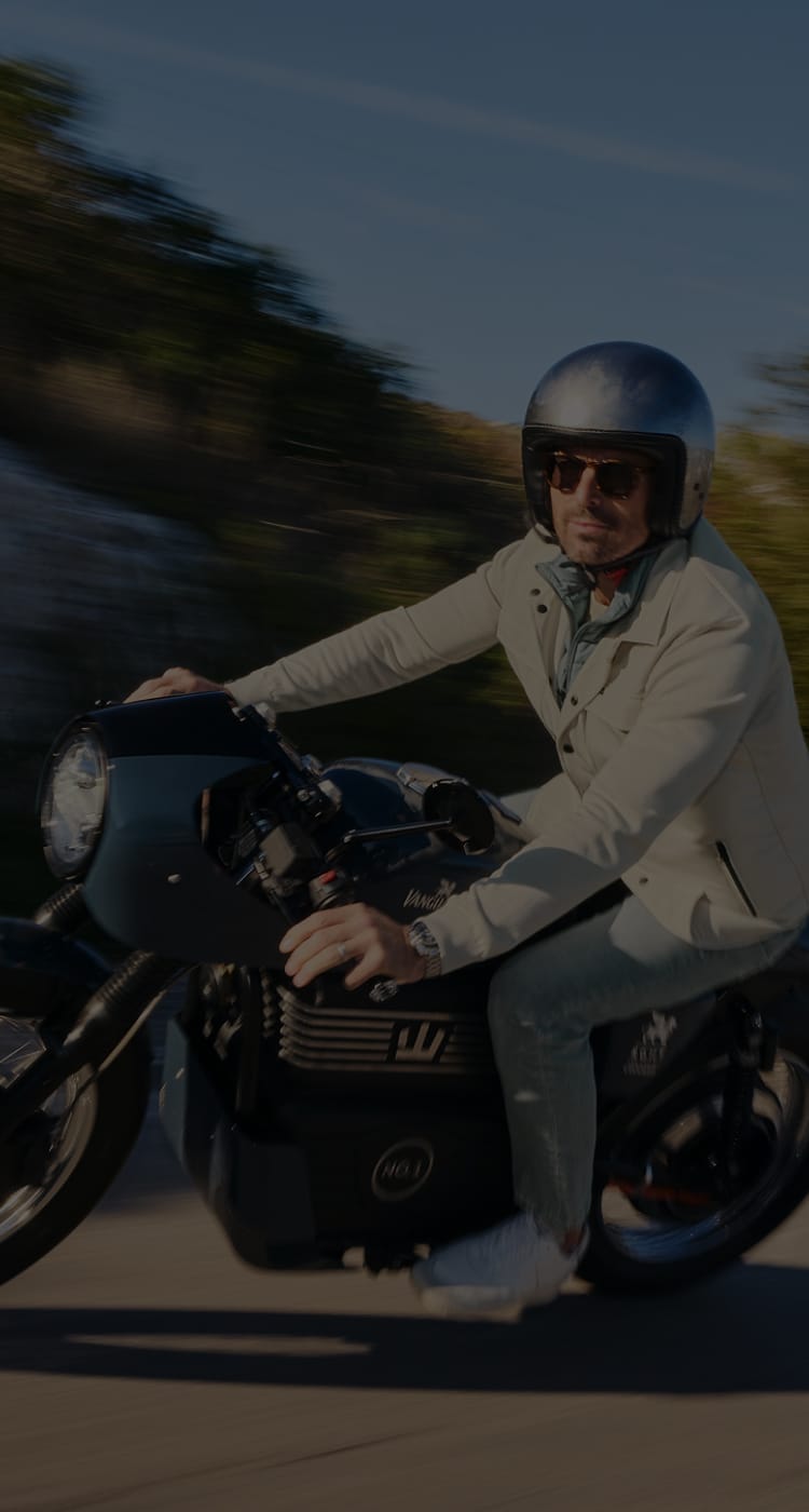 Man in a white jacket and helmet riding a motorcycle on a sunny day, with a blurred background suggesting motion.