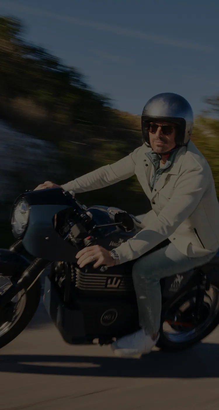 Man in a white jacket and helmet riding a motorcycle on a sunny day, with a blurred background suggesting motion.