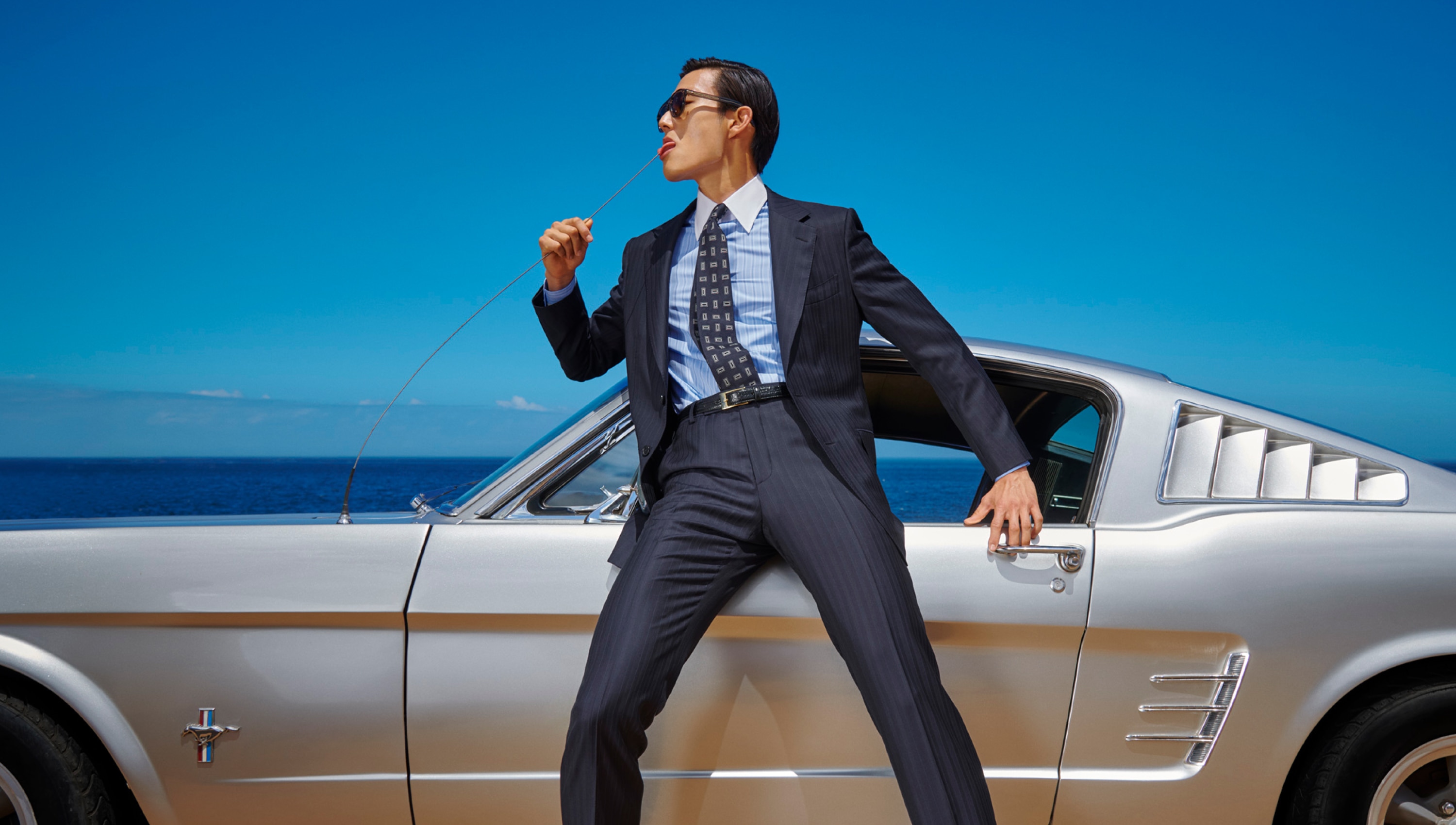 Person in a suit leans on a silver car by the ocean, pulling on a chain. Clear blue sky and sea in the background.
