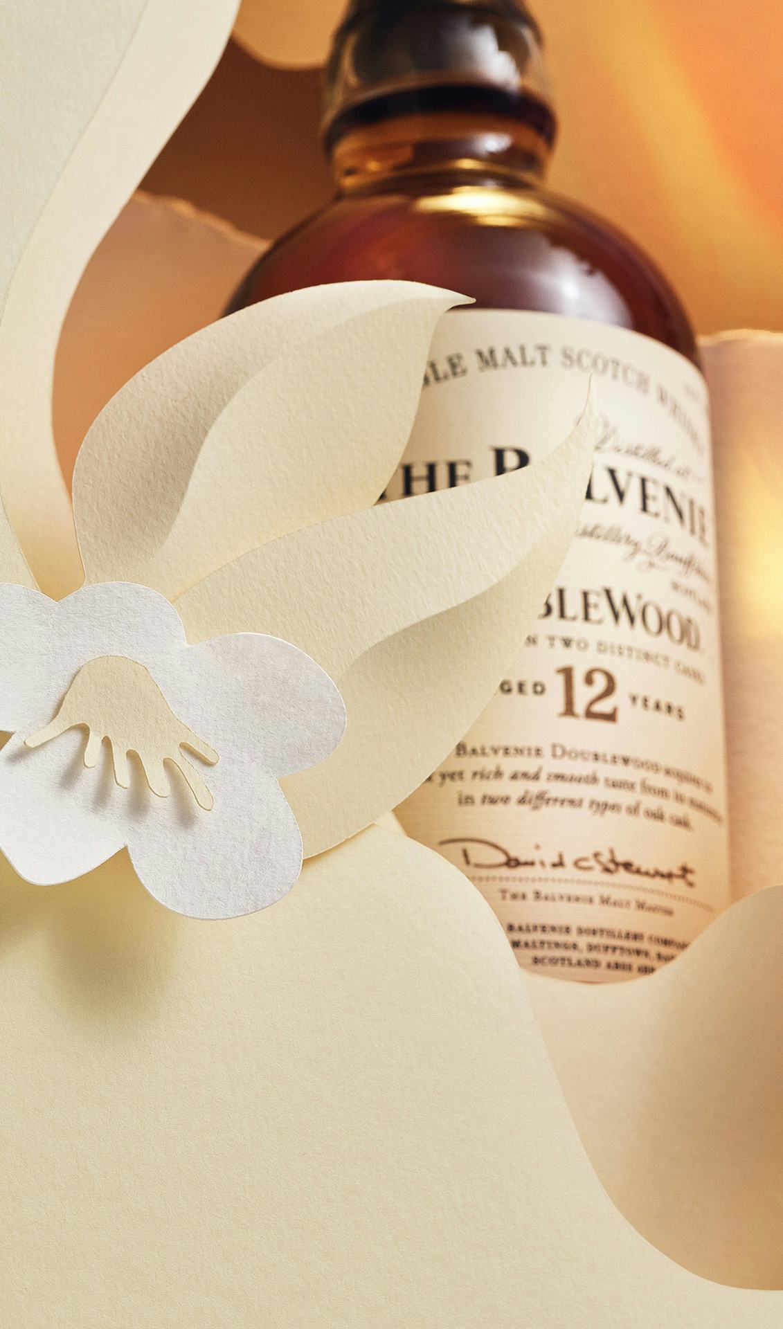 The Balvenie DoubleWood12 in a paper display with golden light and a paper flower