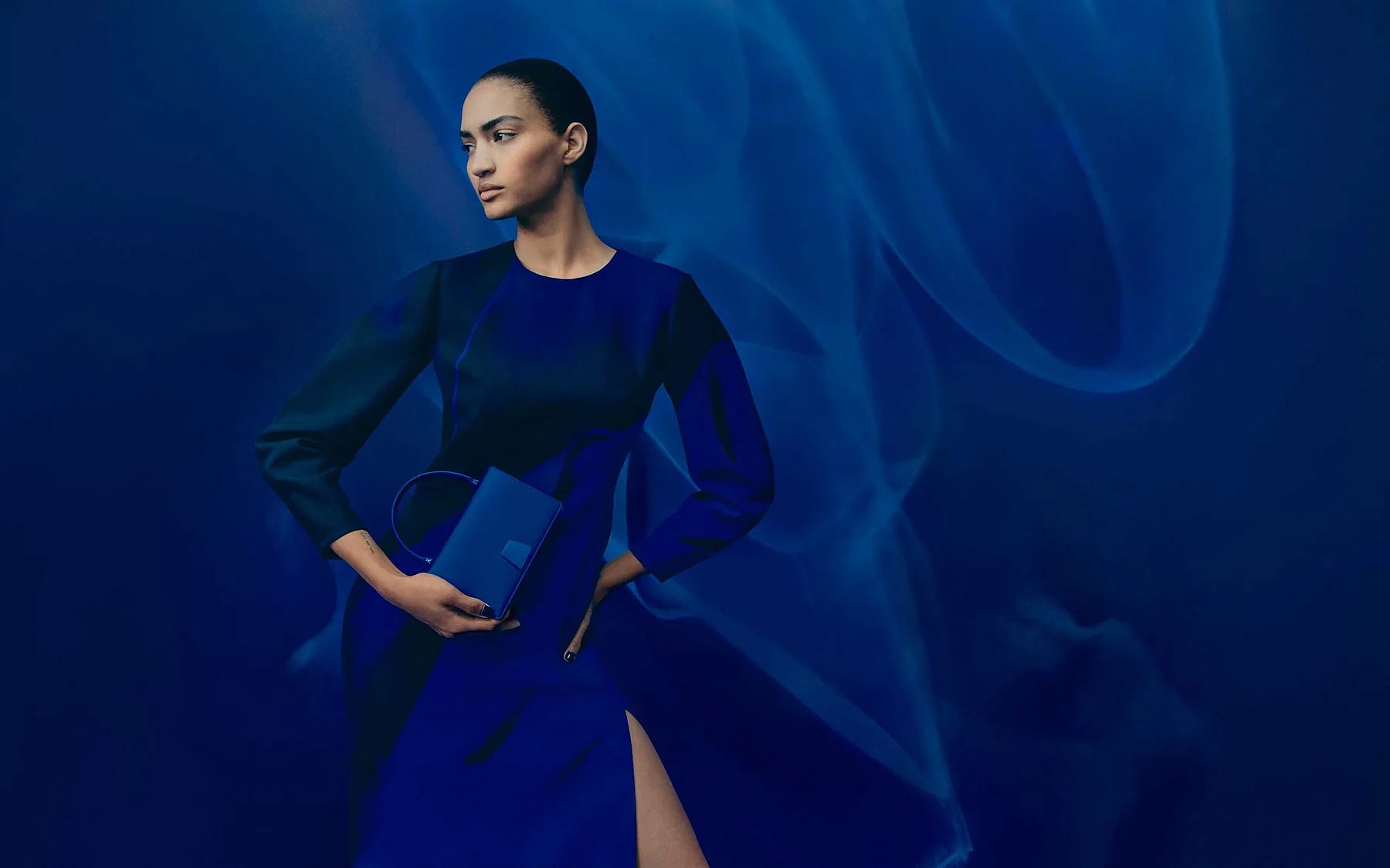A woman in a blue dress poses confidently against a swirling blue background, holding a matching blue handbag.