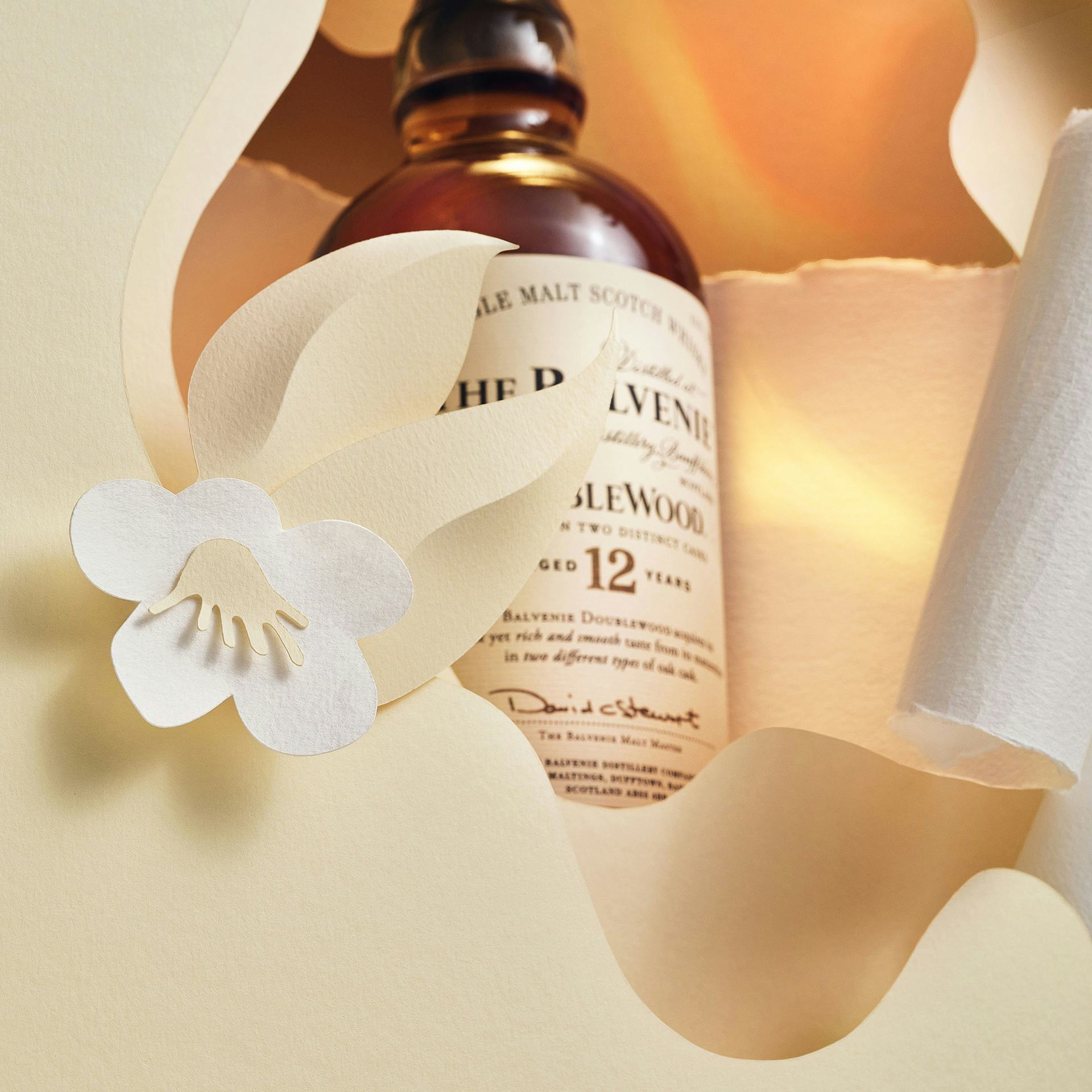 The Balvenie DoubleWood12 in a paper display with golden light and a paper flower