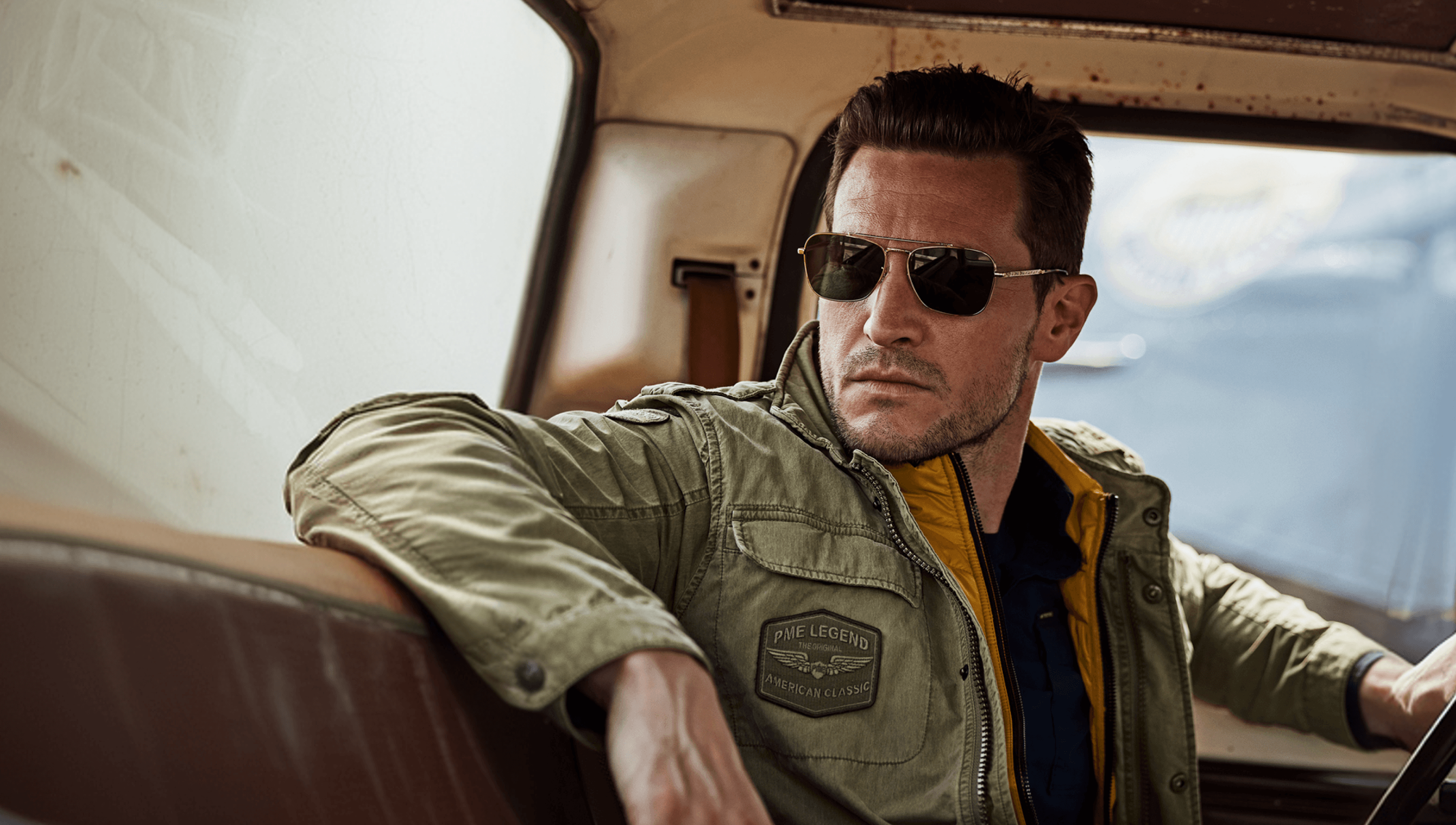 Man in sunglasses and a green jacket sits in a vintage car, looking out the window with a serious expression.