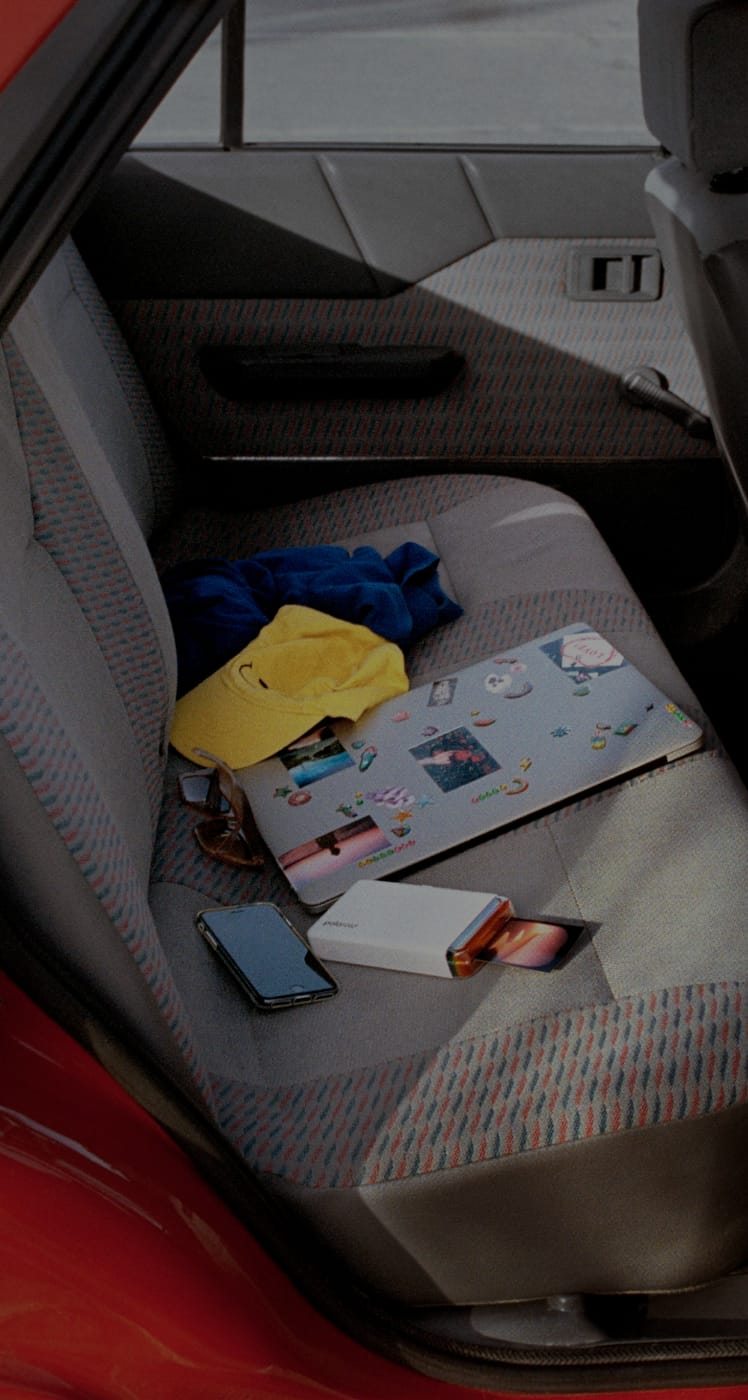 Car backseat with a laptop covered in stickers, a yellow cap, sunglasses, a smartphone, and a polaroid printer on the gray fabric seat.
