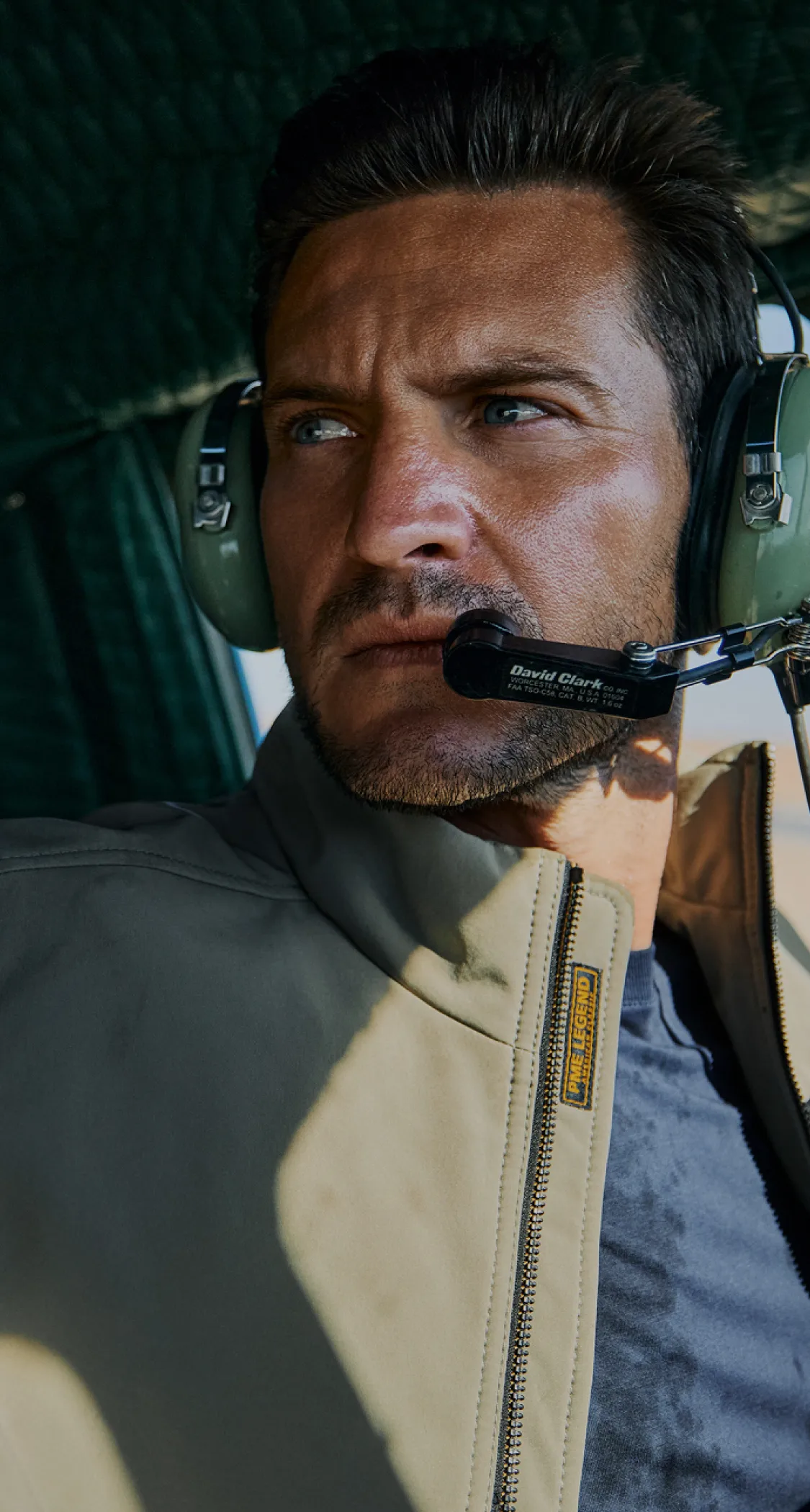 Man wearing a headset with a microphone, looking out of a window. He is dressed in a light jacket and appears focused.